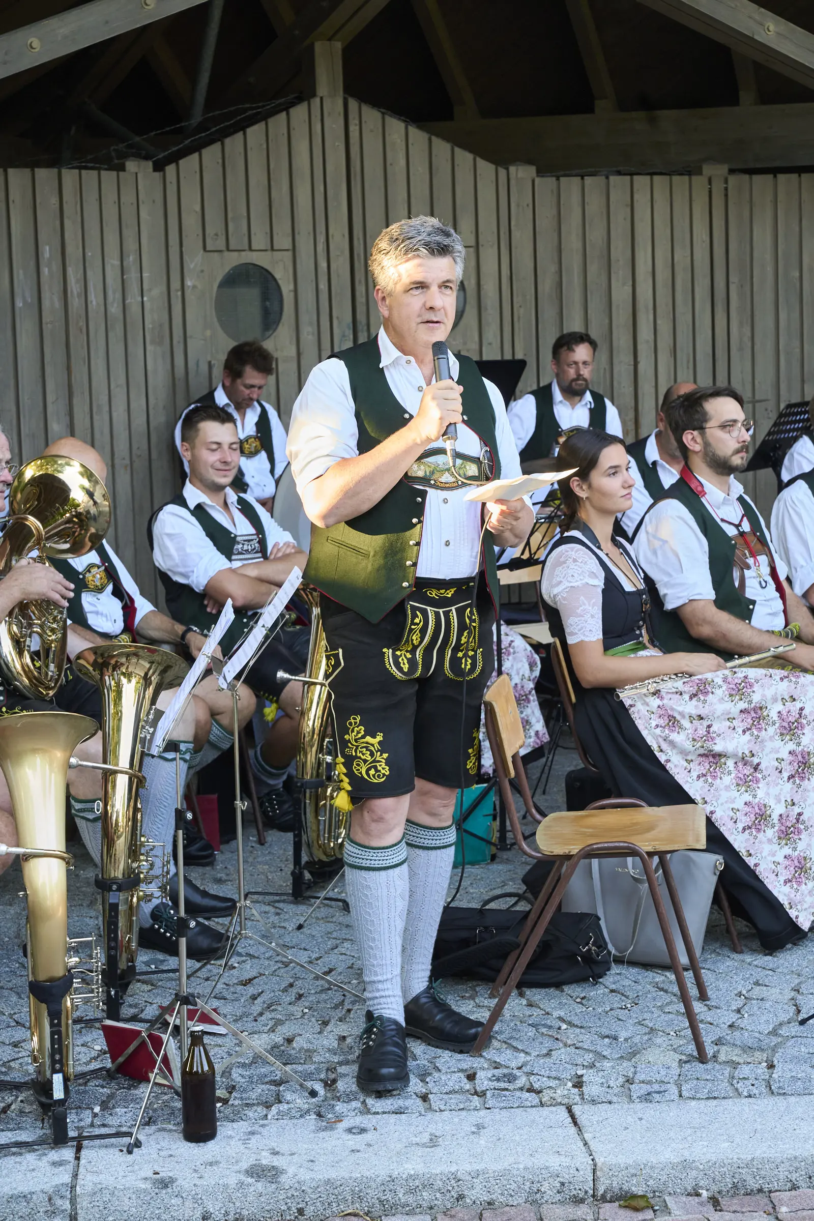Mann in Lederhosen spricht ins Mikrofon, Blasmusik im Hintergrund.